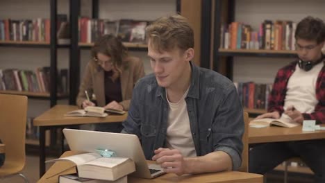 Blonde-caucasian-guy-sitting-in-college-library-and-working-on-laptop,-preparing-for-exams-then-looks-on-a-camera-and-smiling.-His-classmates-and-book-shelves-on-the-background.-Slow-motion