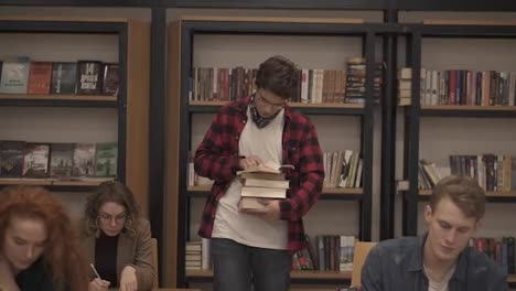 Slow-motion-footage-of-a-guy-in-plaid-shirt-and-headphones-walking-by-row-with-pile-of-books-and-reading.-Other-students-sitting,-studying-in-college,-university-library.-Front-view