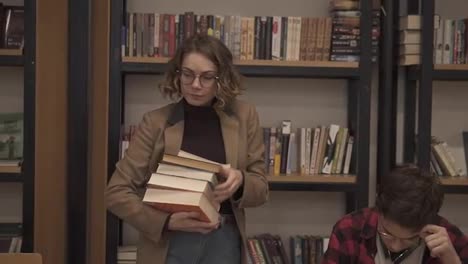 Slow-motion-footage-of-a-girl-in-brown-jacket-and-glasses-walking-by-row-with-pile-of-books-she-took-from-the-shelf-and-starting-to-read-the-book.-Other-students-sitting,-studying-in-college,-university-library.-Front-view