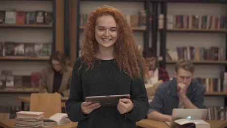 Portrait-of-an-attractive-long-red-haired-european-girl-student-standing-in-high-school-library-smiling-smiling-looking-at-camera-while-holding-a-tablet.-Education,-literature-and-people-concept.-Classmates-on-blurred-background