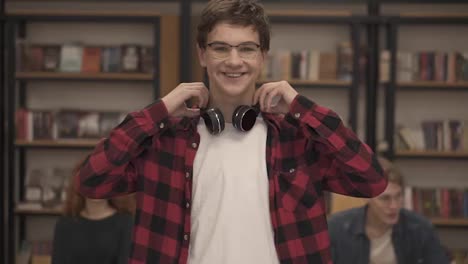 Close-up-portrait-cheerful-young-man-laughing-enjoying-successful-lifestyle-achievement.-European-male-in-plaid-shirt-and-headphones-in-library.-Bookshelf-and-classmates-on-background-college-education
