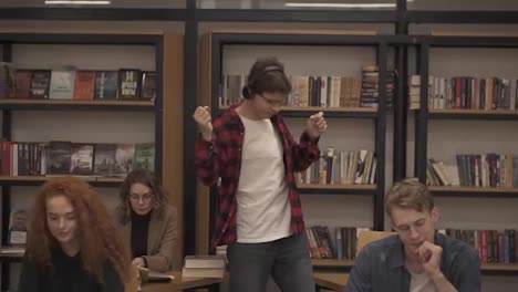 Joyful-excited-handsome-european-man-in-checkered-shirt-performing-expressive-dance-while-listening-groovy-music-in-headphones-in-academic-library-against-bookshelves-background-and-classmates-around