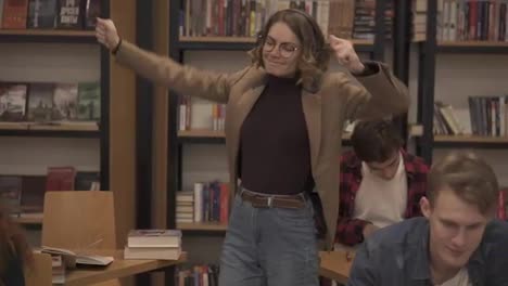 Joyful-excited-european-woman-in-jacket-and-jeans-performing-expressive-dance-while-listening-groovy-music-in-headphones-in-academic-library-against-bookshelves-background-and-classmates-around