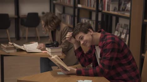 Concentrated-european-male-student-preparing-for-examination-and-reading-books-while-sitting-at-table-at-university-library-with-his-classmates-studying-books