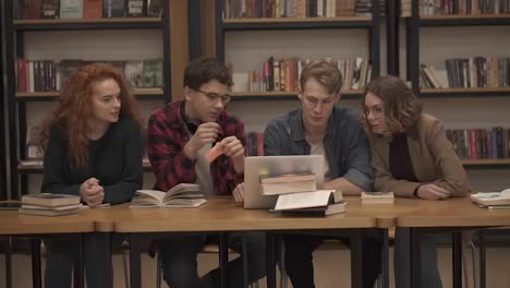 Group-of-young-students-talking-using-laptop-explaining-course-work-study-together-in-library.-Actively-discussing-something-sitting-by-wooden-table.-Slow-motion.-Front-view