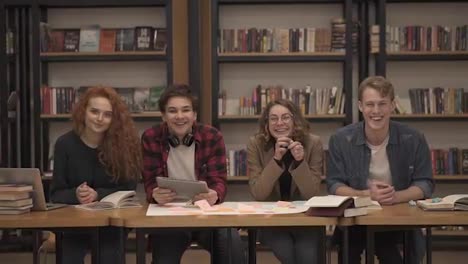 Two-young-male-and-two-female-european-students-sitting-at-the-table-with-books-and-laptop-in-the-library-and-cheerfully-smiling.-Bookshelves-in-library-with-lots-of-books-in-background