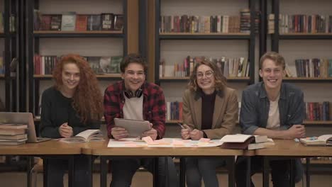 Two-young-male-and-two-female-european-students-sitting-at-the-table-with-books-and-laptop-in-the-library-and-cheerfully-smiling.-Bookshelves-in-library-with-lots-of-books-in-background.-Slow-motion