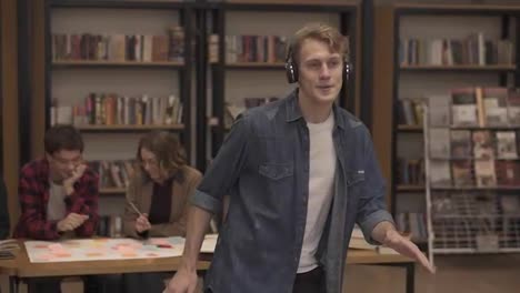 Handsome,-excited-european-man-in-denim-shirt-performing-expressive-dance-while-listening-favourite-music-in-headphones-in-academic-library-against-bookshelves-and-classmates-background.-Slow-motion
