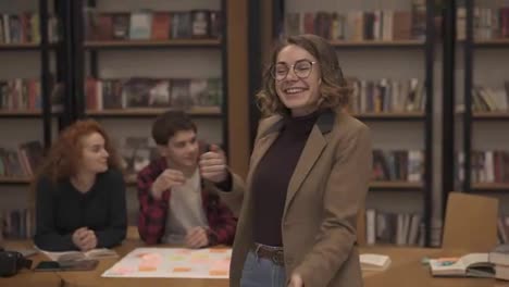 Portrait-of-joyful-excited-european-woman-in-jacket-and-jeans-performing-expressive-dance-while-listening-music-in-headphones-in-academic-library-against-bookshelves-background-and-classmates-around