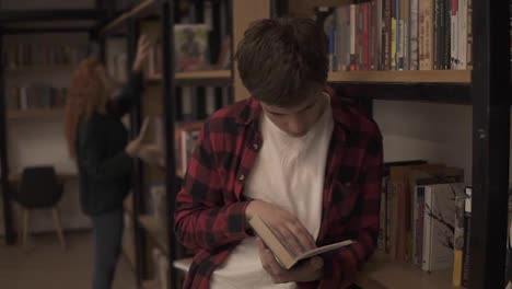 Young-handsome-man-standing-leaning-on-reading-a-book---commuter,-student,-knowledge-concept.-Young-man-in-plaid-shirt-reading-a-book-in-the-college-library