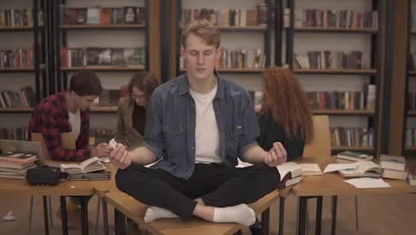 In-the-library-during-the-studying.-Young-male-student-meditating-on-the-desk-in-the-lotos-pose-while-his-classmates-on-the-background-continuing-studying-ignoring-him.-Bookshelves-on-the-background