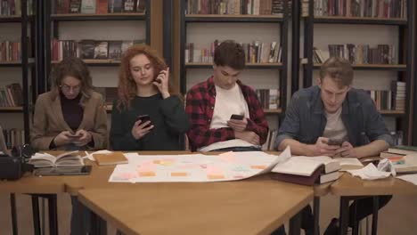 Group-of-four-university-students-two-males,-two-females-sitting-at-the-table-together-during-break-and-using-smartphones---texting,-surfing-internet-while-sitting-in-the-uni-library-back-to-the-bookshelves