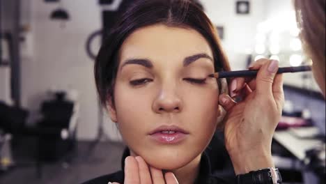 Unrecognizable-make-up-artist's-hand-applies-eyeliner-in-the-eyes'-outer-V-section-of-a-beautiful-attractive-brunette-girl.-Beauty-salon-background.