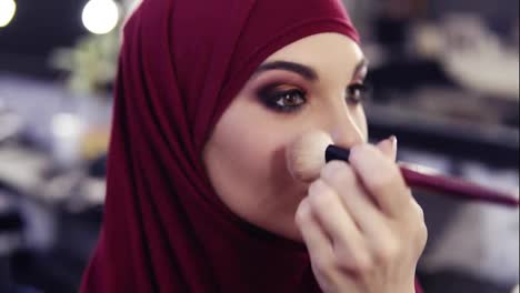 Stunningly-beautiful-girl-with-traditional-arabic-purple-chiffon-hijab-covering-her-head-has-unrecognizable-make-up-artist's-hand-applying-some-blush-on-her-cheeks.