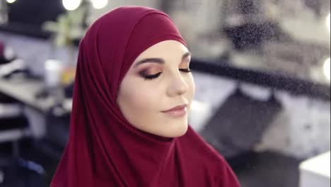 Gorgeous-young-girl-with-hazelnut-eyes-and-purple-hijab-on-her-head-has-make-up-fixating-mist-sprayed-on-her-face-while-getting-final-touches-of-flawless-make-up-look