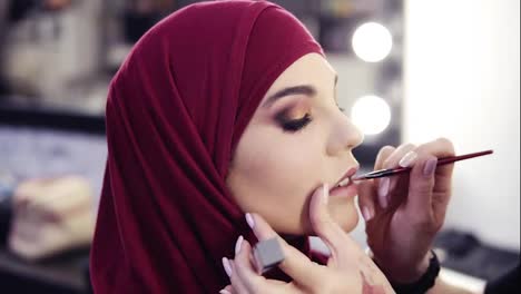 Unbelievably-beautiful-girl-with-hazelnut-eyes-and-purple-hijab-on-her-head-has-unrecognizable-make-up-artist's-hand-accurately-applying-lipstick-with-a-special-brush