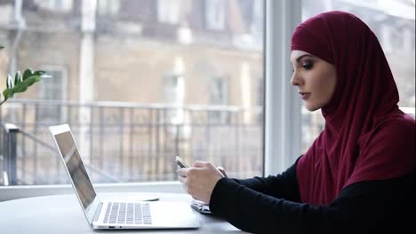 Beautiful-young-female-with-purple-hijab-covering-her-head-scrolls-her-smartphone,-sitting-beside-the-laptop-in-a-light-unrecognizable-indoor-space