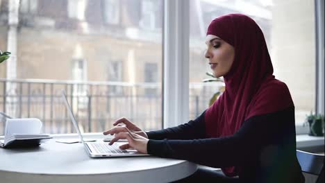 Ein-Junges-Attraktives-Muslimisches-Mädchen-Mit-Hijab,-Das-Ihren-Kopf-Bedeckt,-Tippt-Etwas-Auf-Ihrem-Laptop,-Während-Sie-In-Einem-Angeblichen-Co-Working-Bereich-Mit-Glasfenstern-Im-Hintergrund-Sitzt
