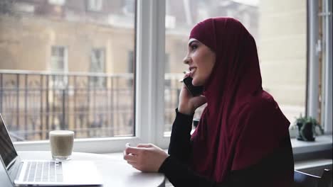 Young-attractive-girl-of-unrecognizable-nationality-with-hijab-on-her-head-is-talking-to-someone-on-the-phone-while-she-has-laptop-and-glass-of-cappuccino-lying-in-front-of-her