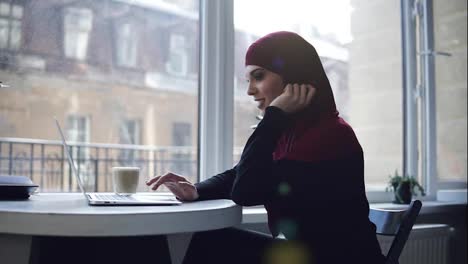 Una-Atractiva-Chica-Musulmana-Con-Hijab-Cubriéndose-La-Cabeza-Mira-Y-Sonríe-Algo-En-La-Pantalla-De-Su-Computadora-Portátil.-Imágenes-En-Cámara-Lenta-En-Interiores