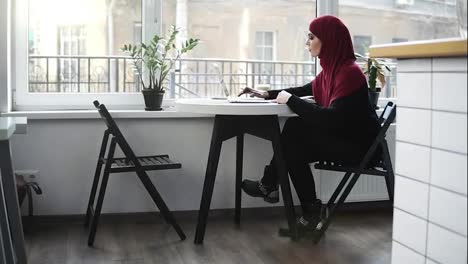Beautiful-muslim-girl-wearing-hijab-is-looking-for-something-on-the-internet-in-her-laptop-and-writes-something-down-while-sitting-in-a-minimalistic-light-place-indoors