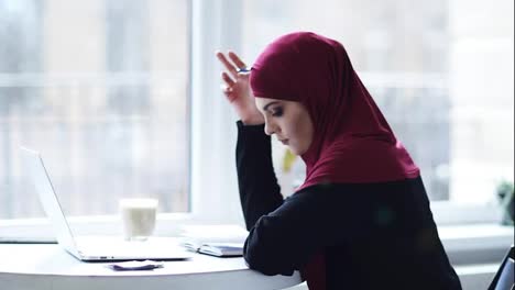 Una-Atractiva-Chica-Musulmana-Está-Escribiendo-Algo-En-Su-Cuaderno-Y-Luego-Comienza-A-Escribir-Algo-En-El-Teclado-De-La-Computadora-Portátil.-Imágenes-En-Cámara-Lenta-En-Interiores
