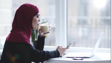 Una-Atractiva-Chica-Musulmana-Con-Hijab-En-La-Cabeza-Está-Bebiendo-Capuchino-Mientras-Busca-Algo-En-Su-Teléfono-Inteligente.-Metraje-En-Interiores