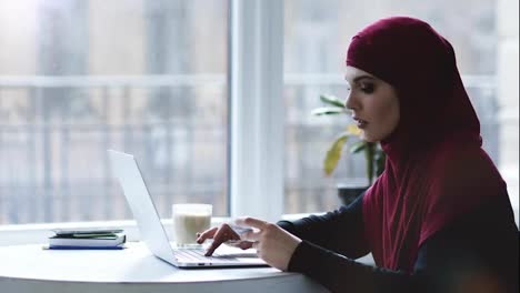 Attractive-muslim-girl-is-typing-some-information-from-a-business-card-on-laptop-keyboard.-Indoors-footage