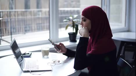 Attraktives-Mädchen-Mit-Traditionellem-Arabischen-Hijab-Arbeitet-An-Etwas-Auf-Ihrem-Laptop-Und-Steckt-Sich-Dann-Kopfhörer-In-Die-Ohren.-Innenaufnahmen-Einer-Wunderschönen-Frau