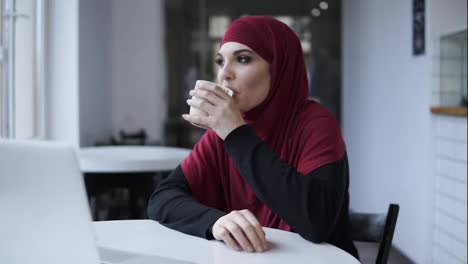 Attraktive-Junge-Schüchterne-Frau-Im-Hijab,-Die-In-Einem-Minimalistischen-Café-Cappuccino-Trinkt.-Innenaufnahmen