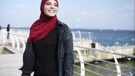 Una-Atractiva-Chica-Musulmana-Con-Hijab-Sonríe-Y-Va-Y-Viene,-Disfrutando-De-Su-Paseo-Junto-Al-Mar.-Actividades-De-Tiempo-Libre.-Metraje-Al-Aire-Libre