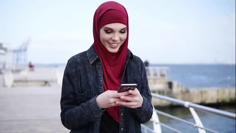 Attraktives-Junges-Mädchen-Mit-Hijab-Auf-Dem-Kopf-Lächelt,-Während-Sie-Jemandem-Eine-SMS-Schreibt-Und-Etwas-Auf-Ihrem-Smartphone-Scrollt.-Zeitlupenaufnahmen-Im-Freien