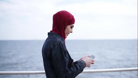 Young-attractive-muslim-girl-with-hijab-on-her-head-is-scrolling-and-typing-something-on-her-smartphone-while-walking-near-the-sea-side
