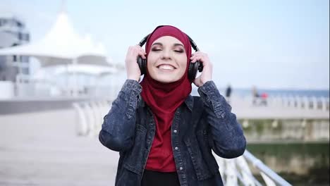 Slow-motion-footage-of-a-young-attractive-girl-with-hijab-on-her-head-putting-headphones-on,-smiling-and-enjoying-the-music-while-walking-near-sea-side