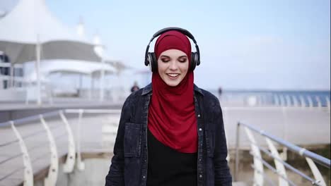 Modern-muslim-girl-with-hijab-covering-her-head-puts-headphones-on-and-starts-walking-somewhere-enjoying-and-dancing-to-the-music