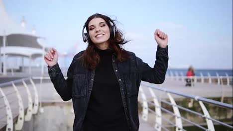 Unbelievably-attractive-young-girl-is-walking-near-the-seaside-with-wireless-headphones-on,-dancing-to-and-enjoying-the-music.-Outdoors-footage