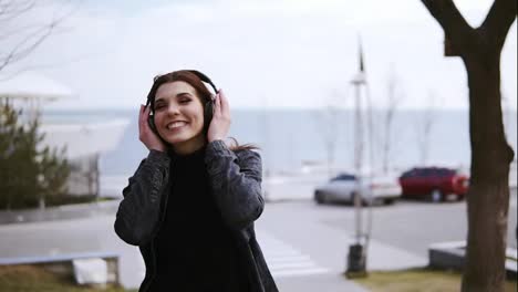 Attractive-young-brunette-girl-with-black-wireless-headphones-on-is-walking-somewhere,-listening-and-singing-to-music.-Enjoying-youth-and-free-time.-Outdoors-footage