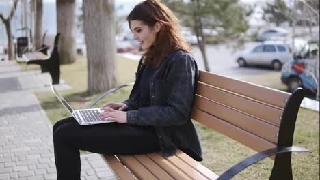 Attractive-female-in-her-20's-wearing-hipster-urban-outfit-sitting-on-the-bench-and-typing-something-on-her-laptop-keyboard