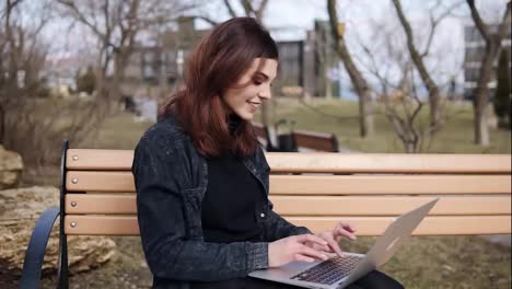 Attractive-female-in-her-20'sis-sitting-on-the-bench-at-park-typing-something-on-her-laptop-keyboard.-Enjoying-free-time.-Outdoors-footage