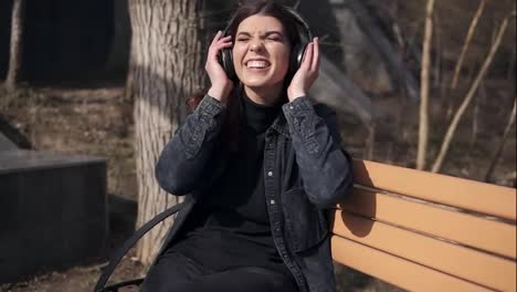 Beautiful-attractive-young-girl-in-black-jeans-jacket-is-sitting-on-the-bench-in-park-enjoying-listening-to-music-in-her-black-wireless-headphones