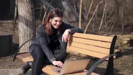 Young-attractive-brunette-girl-is-sitting-on-the-bench-in-park-waving-at-someone-in-her-laptop,-chatting,-laughing-and-having-fun-time.-Outdoors-footage