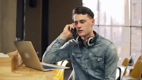 Attractive-male-in-his-20's-in-denim-shirt-with-wireless-headphones-around-his-neck-and-laptop-beside-him,-talking-with-someone-on-the-phone.-Open-co-working-space.-Indoors.