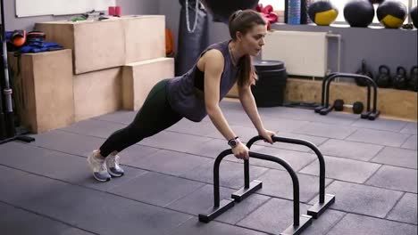 Attractive-sportive-brunette-girl-doing-push-ups-using-push-up-bars-at-the-gym.-Working-on-strong-arms-and-core.-Healthy-lifestyle,-fitness-and-wellbeing.