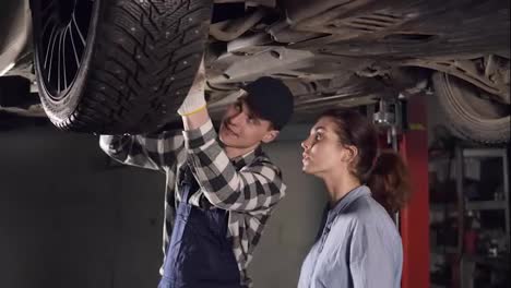 Handsome-mechanic-showing-his-female-client-all-the-problems-in-her-vehicle,-pointing-at-some-mechanisms-on-the-bottom-of-the-car.