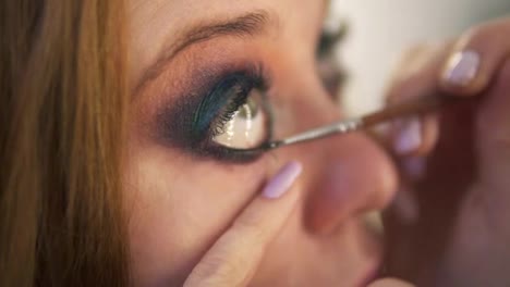 Close-up-of-young-female-model-is-being-maked-up-in-studio.-Artist-is-applying-black-liner-to-lower-lid-of-the-right-eye-using-a-brush.-Side-view