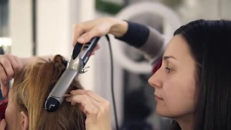 Prepation-process-of-gourgeous-young-woman.-Hairdresser-is-twisting-long-fair-hair-with-a-culring-rod-and-make-up-artist-finishing-visage