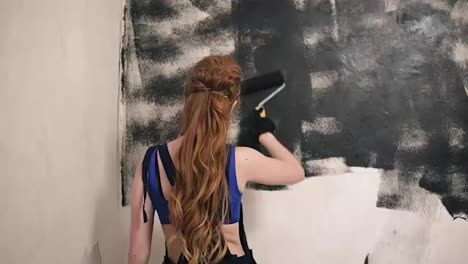 Lovely-woman-in-dark-overall-paints-the-room's-wall-using-a-roller.-Back-view