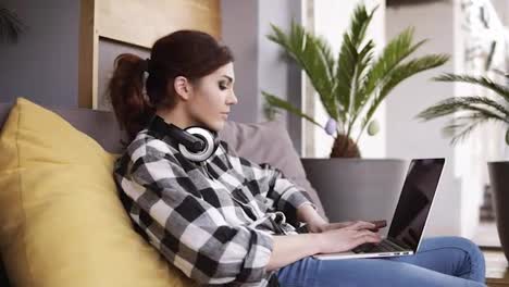 Side-view-of-a-pretty-girl-in-a-plaid-shirt-and-jeans-relaxing-on-a-couch-with-a-laptop-on-legs-and-headphones-on-neck.-Room-with-a-flower