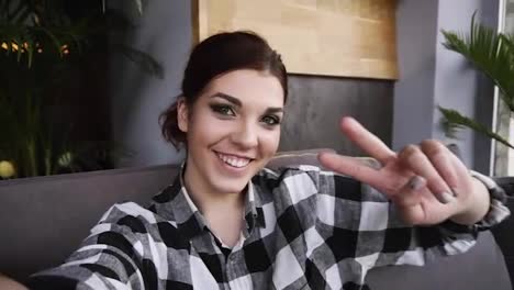 Happy-brunette-girl-dressed-in-plaid-shirt-smiling,-posing,-makes-peace-signs-with-fingers-making-self-portrait---point-of-view-of-camera.-Slow-motion
