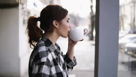 Gorgeous-girls-view-standing-near-the-windows.-Drinking-a-tea-with-a-white-cup.-Side-view.-Blurred-background